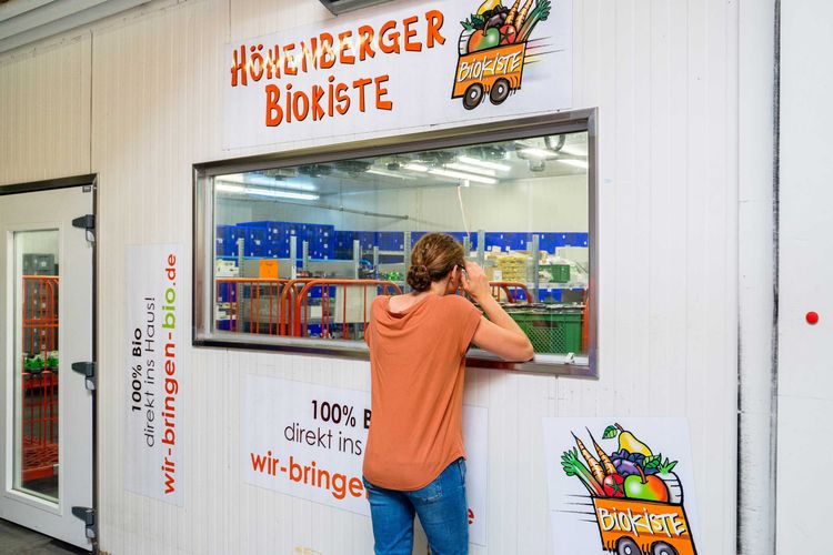 KI generiert: Ein Mann schaut durch ein Fenster in einen Raum mit Lebensmitteln bei der "Höhenberger Biokiste". Auf dem Schild steht, dass es sich um einen Lieferdienst für Bio-Produkte handelt.